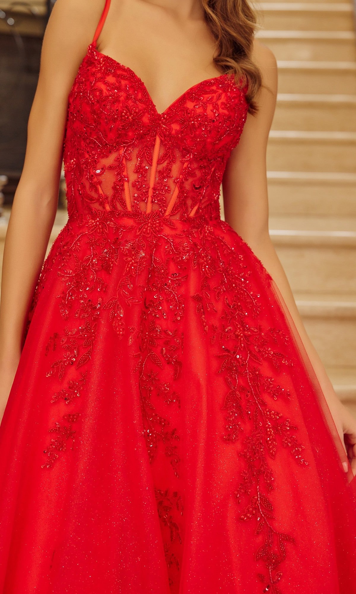 A woman wearing the Nox Anabel H1883 long prom dress in red, featuring a beaded corset bodice and embroidered floral appliques, stands before a staircase.